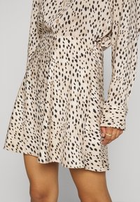 Světle béžové šaty s černým skvrnitým vzorem, dlouhými rukávy a volným střihem. Měkká textura tkaniny s rozšířeným lemem.