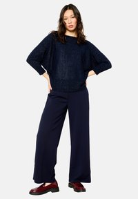 Navy knitted sweater with sparkly texture, wide sleeves; paired with high-waisted, dark navy trousers and burgundy patent loafers.