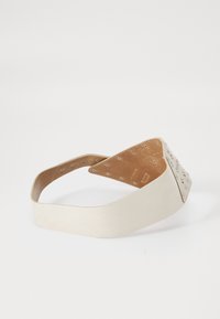 Cinturón ancho de cuero beige con múltiples ojales de metal y una pieza triangular en el extremo, dispuesto sobre una superficie blanca.