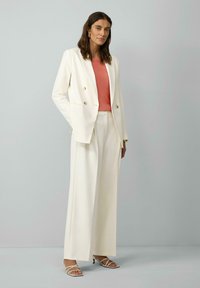 Biała dwurzędowa marynarka, koralowa top o fakturze i szerokie spodnie. Sandały mają cienkie paski. Gładki materiał, dopasowany krój.