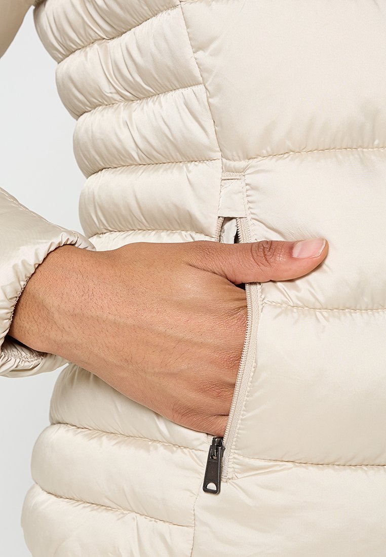 Giacca puffer beige con design trapuntato, dotata di tasca con zip e tessuto testurizzato. Mano appoggiata sulla tasca.