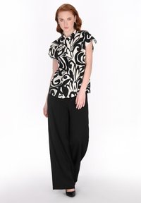 Schwarz-weiße gemusterte Kurzarmbluse; kombiniert mit weitgeschnittenen schwarzen Hosen; glattes Material; klassischer Kragen; schlichte schwarze Pumps.