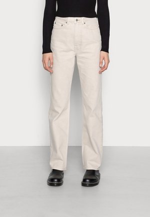 Person trägt cremefarbene Jeans mit geradem Bein, schwarzen gerippten Langarmtop und schwarze Leder-Stiefeletten, steht auf grauem Boden.