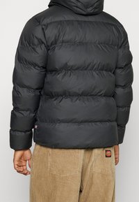 Black puffer jacket with horizontal quilted sections, featuring a hood and elastic cuffs. Worn over light brown corduroy pants.