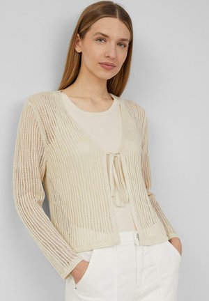 Beige gebreide cardigan met een textuur patroon, open voorkant en striksluiting. Draagt over een lichtbeige top, gecombineerd met witte broek.