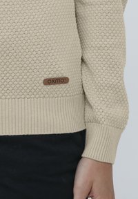 Beige neulepaita, jossa on teksturoitu mehiläispesäkuvio, ribbireunat ja ruskea nahkalappu, jossa on teksti "OXMO" alataskussa.
