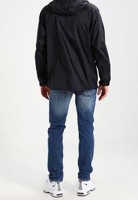 Giacca nera leggera con cappuccio, abbinata a jeans in denim blu e scarpe da ginnastica bianche con un accento grigio.