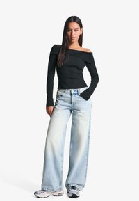 Top noir à côtes, décolleté épaule émoussée, associé à un jean large bleu clair. Le modèle porte des sneakers gris et noir, affichant une tenue décontractée et relax.