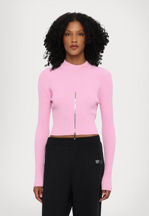 Sopiva, ribatty vaaleanpunainen crop-top, jossa on korkea kaula-aukko ja hopeinen vetoketju, yhdistettynä mustiin, löysiin housuihin, joissa on hienovarainen embleemi.