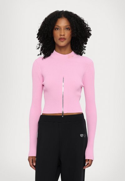 Tett, ribbet rosa crop-top med høy hals og sølvglidelåsdetalj, kombinert med svarte, løstsittende bukser med et subtilt emblem.