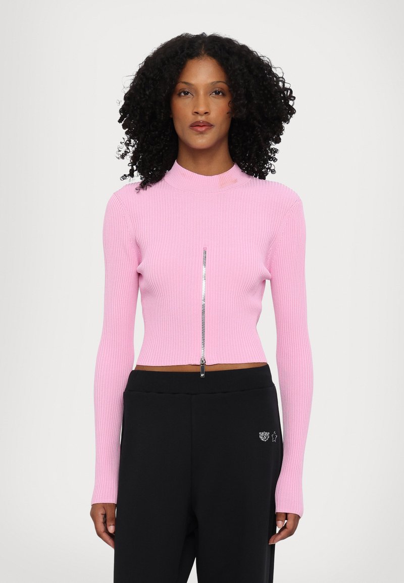 Feszes, bordás rózsaszín crop top magas nyakkal és ezüst cipzáros részletekkel, fekete, laza szabású nadrággal párosítva, amelyen egy diszkrét embléma található.