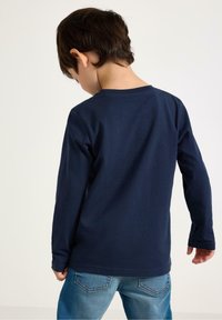 Top a maniche lunghe di colore blu navy in tessuto a costine, con scollatura rotonda e orlo dritto, abbinato a jeans in denim. Vista posteriore del bambino.