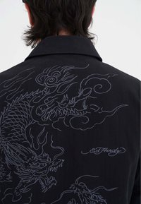 Czarna kurtka z bardzo szczegółową szaro-skalną haftowaną smoczą aplikacją oraz podpisem "Ed Hardy" na plecach, noszona przez osobę zwróconą tyłem do kamery.