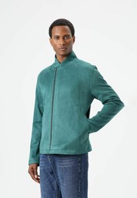 Turkooise suède jas met een hoge kraag en een rits aan de voorkant, met een gladde textuur en een aansluitend ontwerp. Gedragen over een spijkerbroek.