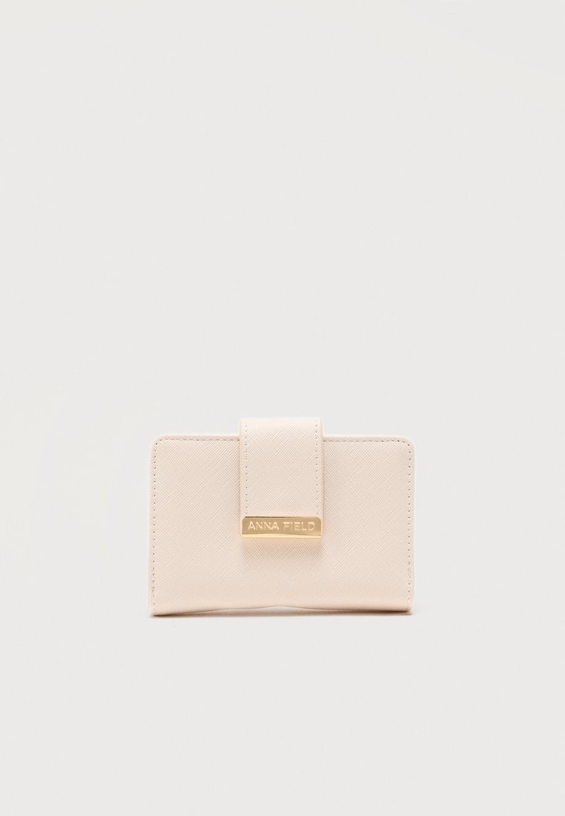 Συμπαγές μπεζ πορτοφόλι με υφή στην επιφάνεια και μπροστινό λουράκι με χρυσή μεταλλική πλάκα με χαραγμένο το "ANNA FIELD".