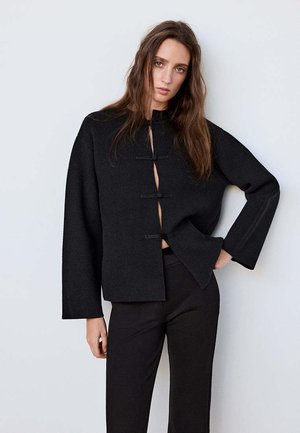 Femme aux longs cheveux bruns porte une veste noire ample avec des boutons à bascule et un pantalon noir, debout devant un fond clair uni.