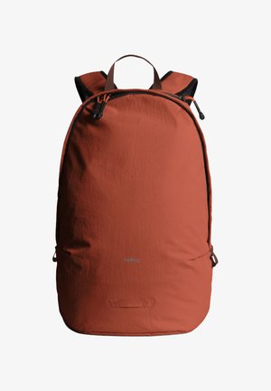 Zaino color ruggine con spallacci imbottiti neri, chiusura con doppia cerniera, maniglia per il trasporto e logo frontale discreto su sfondo bianco.