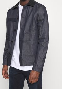 Denim jacket in dark blue with a black collar, button front, chest pocket, and side pockets; smooth texture and tailored fit.
