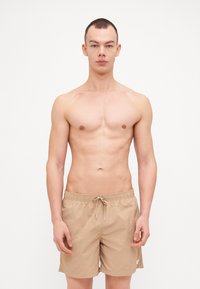 Beige fürdőnadrág elasztikus derékkal és zsinórral. Könnyű anyag, oldalsó zsebekkel és minimális logózással.