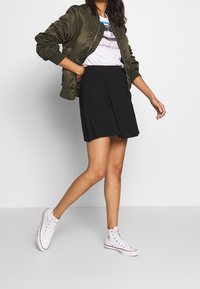 Giacca bomber verde oliva, t-shirt grafica bianca, gonna nera svasata e sneakers alte bianche. Tessuto morbido, vestibilità rilassata.