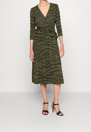 Femme portant une robe portefeuille vert olive avec des rayures zébrées foncées, manches trois-quarts, longueur midi et sandales à talons noirs.