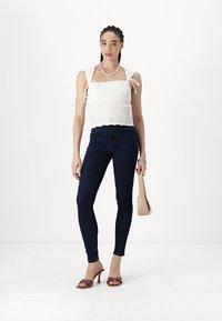 White smocked crop top with tie straps, paired with dark blue skinny jeans and brown heeled sandals, holding a beige handbag.