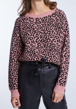 Nainen, joka pukeutunut vaaleanpunaiseen ja mustaan leopardikuvioiseen villapaitaan, joka on sullottu korkeavyötäröisten mustien nahkahousujen sisään, joissa on sidottava vyö.