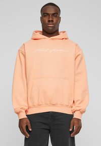 Sudadera color durazno con un bolsillo en la parte frontal, puños y dobladillo acanalados. Presenta texto bordado en blanco en el pecho. Textura suave, similar al algodón.