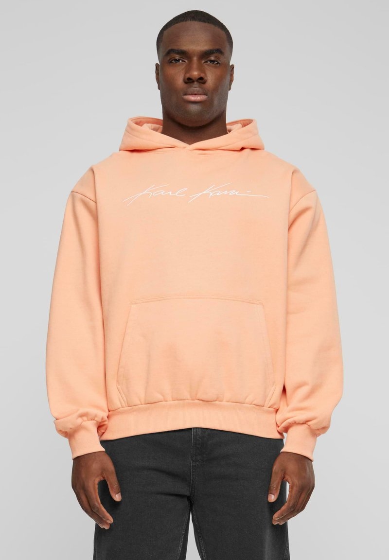 Sudadera color durazno con un bolsillo en la parte frontal, puños y dobladillo acanalados. Presenta texto bordado en blanco en el pecho. Textura suave, similar al algodón.