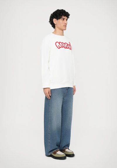 Sudadera blanca con gráfico rojo "MARWI", combinada con jeans de corte amplio en azul. El modelo lleva zapatillas oliva y negras. Fondo liso.