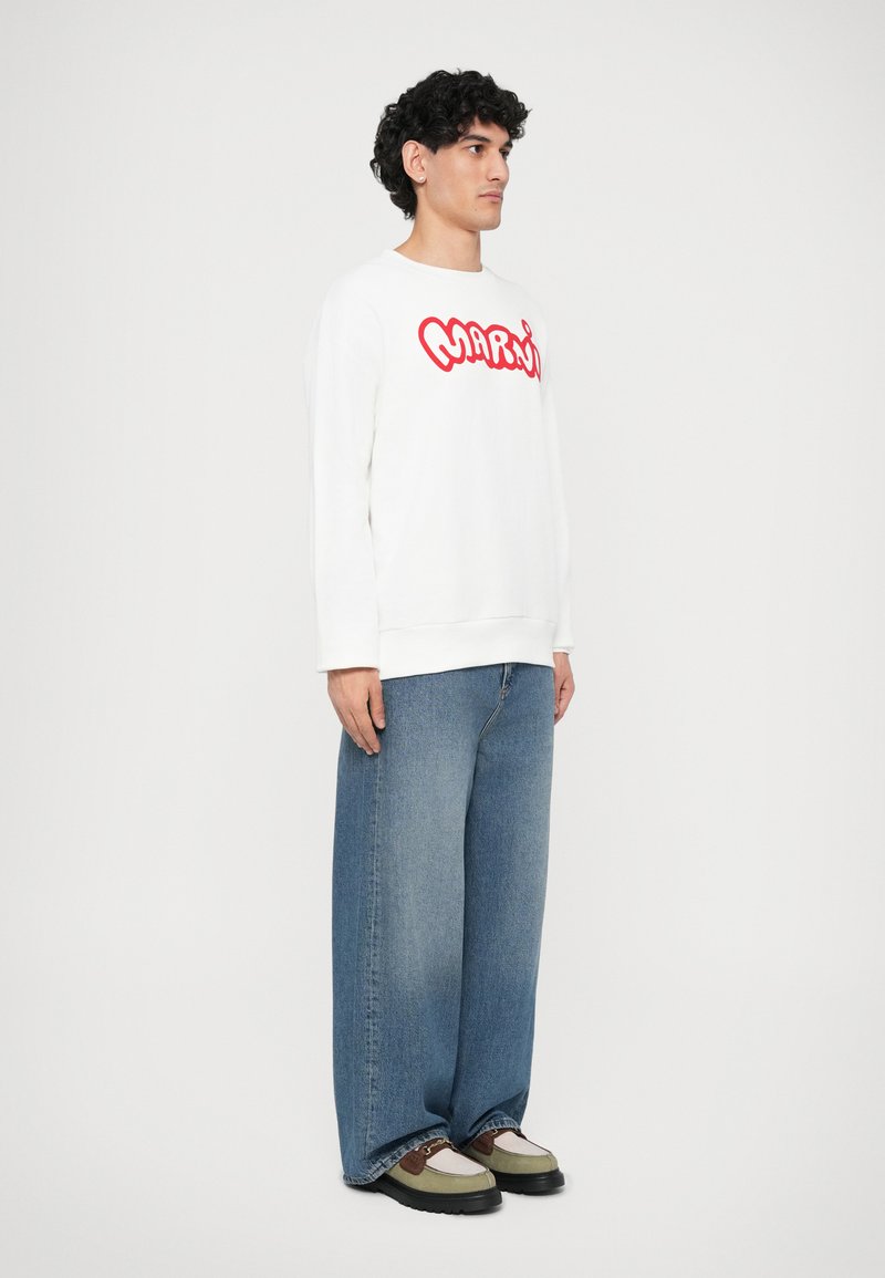 Vit sweatshirt med röd "MARWI"-grafik, tillsammans med löst sittande blå jeans. Modellen bär olivgröna och svarta sneakers. Enkelt bakgrund.