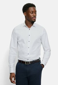 Weißes Langarmhemd mit Kentkragen, ausgestattet mit blauen Knopfdetails und einer glatten Textur, kombiniert mit dunklen Hosen und einem braunen Gürtel.