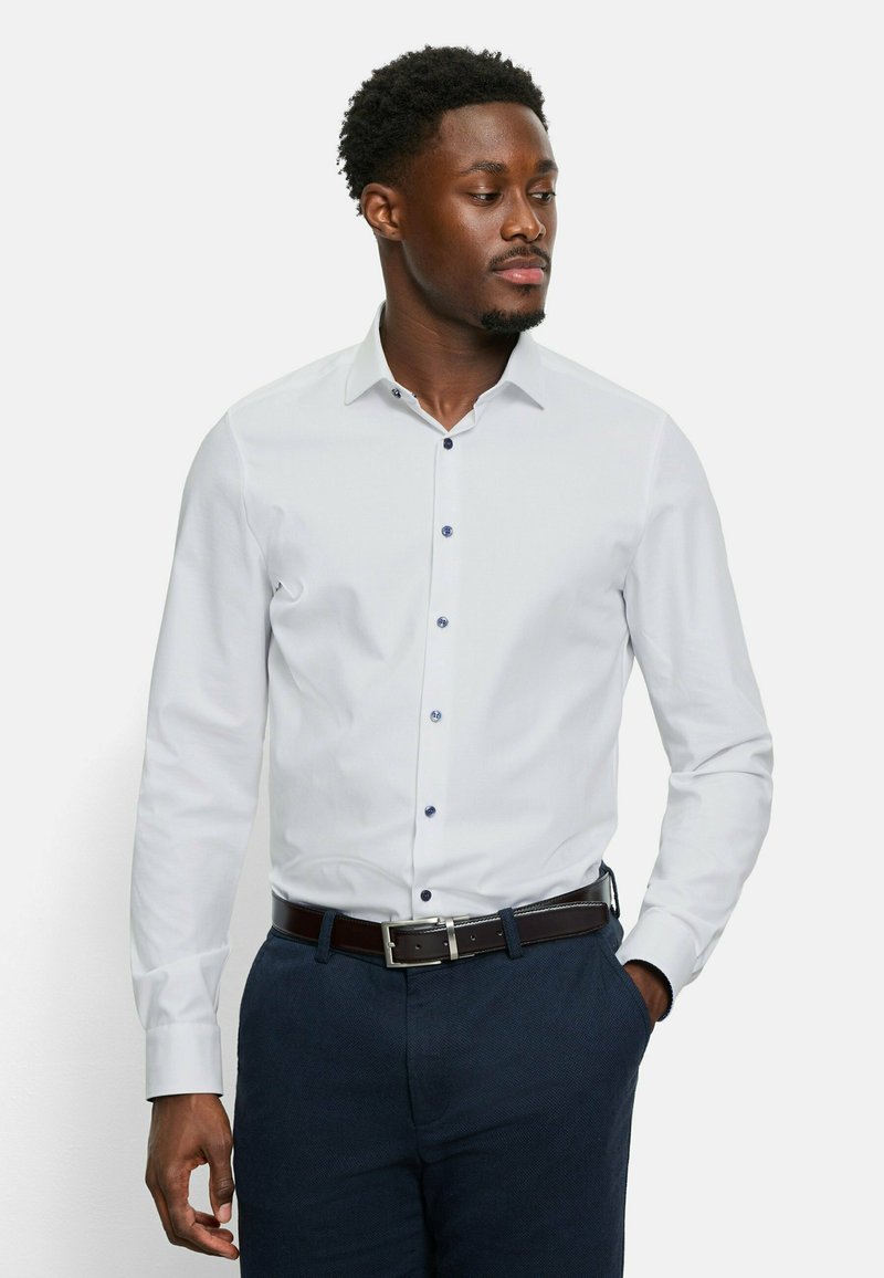 Weißes Langarmhemd mit Kentkragen, ausgestattet mit blauen Knopfdetails und einer glatten Textur, kombiniert mit dunklen Hosen und einem braunen Gürtel.