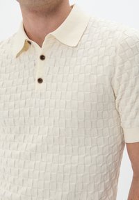 Rövid ujjú krémszínű poloing, texturált kötött anyagból készült, rácsos mintával, három barna gombbal és klasszikus gallérral.