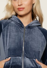 Veste à capuche zippée en velours bleu foncé. Présente une texture douce, des poches latérales et une fermeture éclair lisse. Conception légère et décontractée.