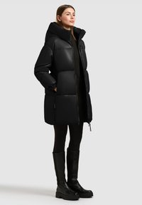 Zwarte pufferjas met een hoge kraag, voorzien van een glanzende afwerking, horizontale doorgestikte structuur, ritssluiting en zijzakken. Accenten met trekkoorden.