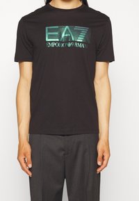 Svart bomullströja med en grafisk "EA7 EMPORIO ARMANI"-logotyp i turkos. Korta ärmar, rundad hals, slim fit-design.