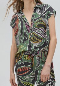 Camicia a maniche corte con bottoni di colore verde, decorata con motivi di foglie colorate in rosso, rosa e giallo. Tessuto liscio, vestibilità comoda.