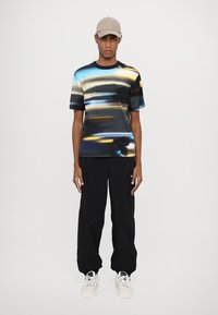 Short-sleeve, black t-shirt featuring a colorful, abstract design; paired with loose black pants and white sneakers. Cap is beige.