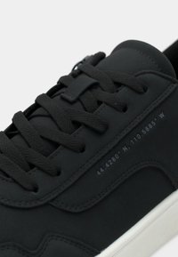 Μαύρο αθλητικό παπούτσι με δερμάτινο επάνω μέρος με υφή, επίπεδα μαύρα κορδόνια και λευκή καουτσούκ σόλα. Διαθέτει ανάγλυφα συντεταγμένες στο πλάι.