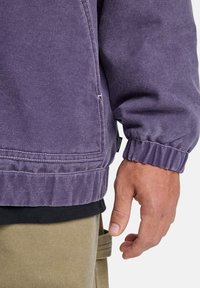 Purple jacket with a textured finish, elastic cuffs, and white stitching, layered over a black shirt and khaki pants.