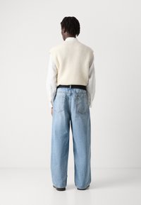 Chaleco crema de gran tamaño sobre una camisa blanca, combinado con jeans de pierna ancha de color azul claro. Las características destacadas incluyen una cinturilla negra y dos bolsillos traseros.