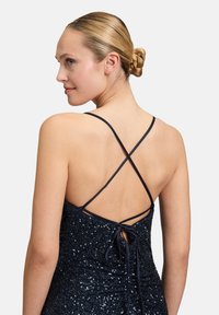 Ein marineblaues Paillettenkleid mit tailliertem Schnitt, kreuzgeschlungenen Rückenträgern und einem gebundenen Verschluss, das die schimmernde Textur hervorhebt.