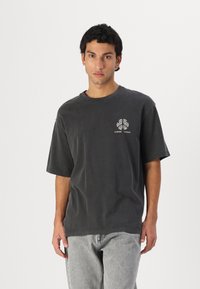 Šedé oversize tričko vyrobené z bavlny, s kulatým výstřihem a krátkými rukávy. Výrazný potisk loga na levé části hrudi. Minimalistický design.