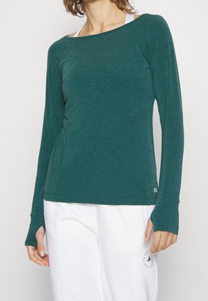 Nainen pukeutuneena teeltiin siniseen pitkähihapaidalle, jossa on peukalonreiät, ja valkoisiin housuihin, joissa on musta ympyrälogo reisiosassa.