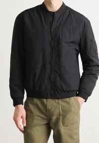 Chaqueta bomber negra con cuello alto, cierre de cremallera, puños de canalé y un bolsillo en la manga izquierda; presenta una textura suave y ligera.