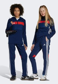 Chándales azul marino con acentos rojos, que incluyen una sudadera con cremallera y pantalones largos, ambos con rayas blancas. Una persona sostiene zapatos de fútbol negros.
