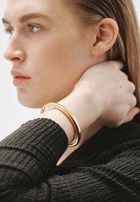 Pulsera de metal dorado en forma de bangle descansando sobre una funda negra texturizada. Diseño suave y redondo con una superficie reflectante.