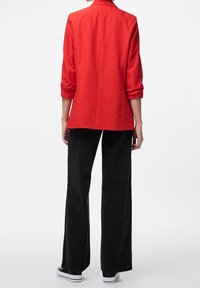 Veste blazer rouge aux manches retroussées, coupe ajustée et fente au dos, associée à un pantalon noir à jambes larges et des baskets blanches. Tissu lisse et structuré.