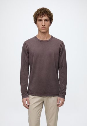 Jeune homme aux cheveux bouclés portant une chemise marron foncé à manches longues et un pantalon beige, debout devant un fond blanc uni.